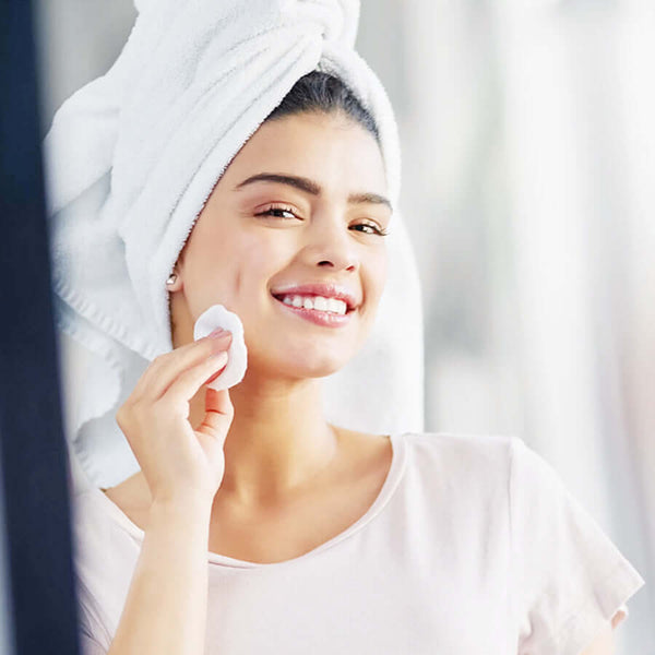 Woman with a towel on her head using a reusable cotton makeup remover pad, smiling and enjoying her skincare routine.
