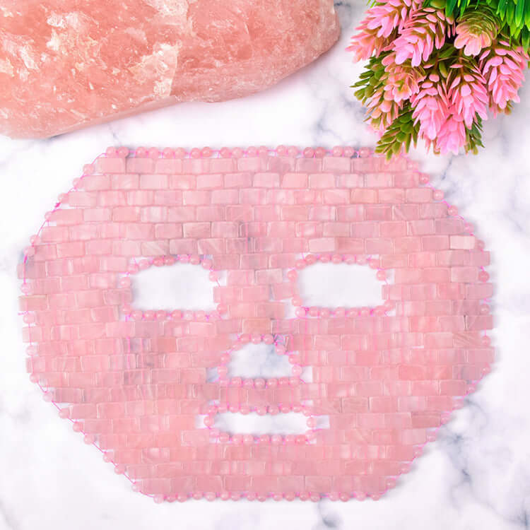 Jade face mask made of pink jade pieces on a marble background with flowers, ideal for facial massage treatment.